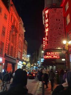 people are walking on the sidewalk in front of tables dancing sign at night, with cars parked along the street