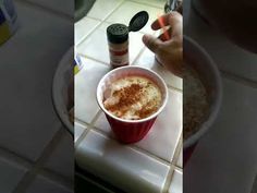 a person holding a spoon over a cup filled with liquid and cinnamon on top of a white tile counter