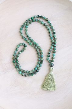 This mala necklace is made with 6-7mm African turquoise beads and a 10mm African turquoise guru bead. The beads are a green turquoise color with a brownish black matrix. Each strand of beads is unique. If you would like a photo of your mala before it is shipped out, please feel free to message me.  This mala is hand-knotted and made in the traditional way with 108 beads. It is 32" in length and the sage green tassel is available as either 2" or 3" long. The tassel is handmade using silk thread.  African turquoise encourages positive change, optimism and balance. It opens the mind to new ideas and helps us confidently walk towards our dreams. African turquoise is part of the jasper family. This mala will come in a stamped cotton bag and box with a card explaining the gemstone meaning. What Green Gemstone Beads Mala For Healing, Spiritual Jade Beaded Necklaces For Meditation, Green Round Beads Spiritual Crystal Necklace, Green 8mm Beads Mala Gift, Green Gemstone Beads Mala As Gift, Turquoise Spiritual Beads For Meditation, Spiritual Turquoise Beads For Meditation, Green Crystal Necklaces With Round Hand-strung Beads, Adjustable Spiritual Turquoise Necklace For Meditation