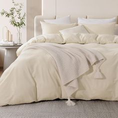 a bed with white linens and pillows in a neutral room, next to a plant