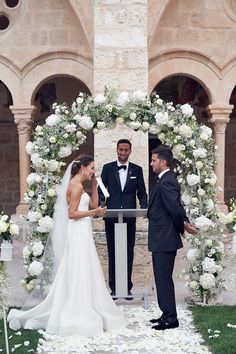 a bride and groom standing in front of an arch with white flowers on the grass