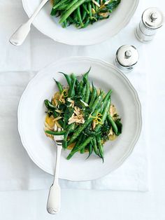 two white plates topped with green beans and noodles next to silverware on a table