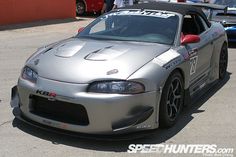 a silver sports car parked in a parking lot with other cars behind it and people standing around