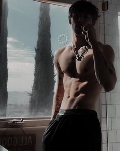 a shirtless man standing in front of a window talking on a cell phone while holding his hand up to his ear