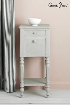 a white table with a bowl on it next to a striped curtain and pink wall