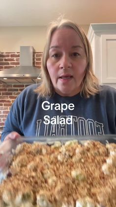 a woman holding up a tray of food in front of her face with the words grape salad on it