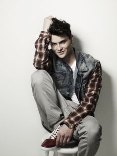 a young man sitting on top of a stool wearing a jean jacket and sneakers with his hands behind his head