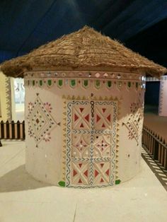 a small round structure with a thatched roof and decorative designs on the sides, in front of a fence