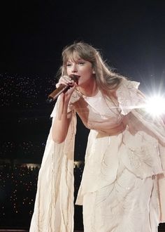 a woman in a white dress holding a microphone and standing on stage with lights behind her