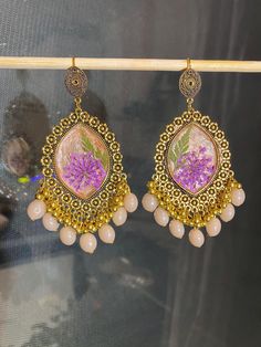 These handmade resin earrings feature a vibrant purple flower encased in a beige background, complemented by gold-earring and dangling beads. Perfect for adding a boho-chic touch to any outfit. Bohemian Gold Resin Earrings, Purple Traditional Earrings For Gift, Pink Bohemian Jewelry With Pressed Flowers, Bohemian Pink Jewelry With Pressed Flowers, Handmade Purple Earrings For Festive Occasions, Bohemian Style Resin Flower Earrings As Gift, Unique Flower-shaped Resin Earrings, Festive Handmade Purple Earrings, Dangle Earrings With Flower Charm In Resin