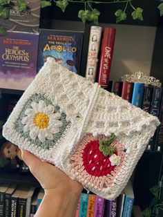 a hand holding up a crocheted box with a strawberry on it in front of bookshelves