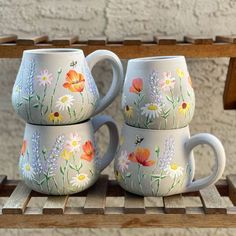 three mugs with painted flowers on them are sitting on a wooden shelf next to a brick wall