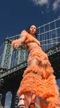 a woman in an orange feathered dress is standing near a bridge and looking up at the sky