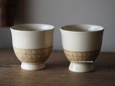 two white cups sitting on top of a wooden table next to each other with gold trimmings