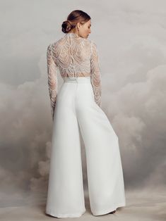 a woman wearing white pants and a sheer top with an open back, standing in front of clouds