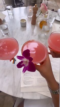 two people holding up pink drinks at a table with flowers in their glasses and menus on the table