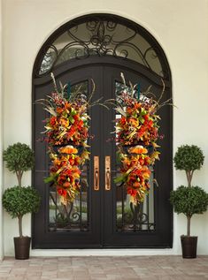 two large wreaths on the front door of a house with potted trees and bushes