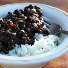 a white bowl filled with rice and meat on top of a wooden table next to a fork