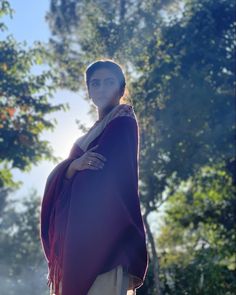 a woman wearing a purple shawl standing in front of trees with her hands on her hips