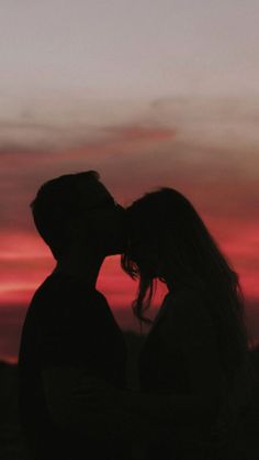 a man and woman kissing in front of a red sky at sunset with the sun setting behind them