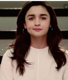 a woman with long dark hair and large hoop earrings looks at the camera while standing in front of stairs