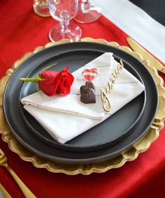 a black plate topped with a red rose next to a white napkin and gold trim