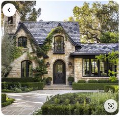 a stone house with black shutters and windows in the front yard is surrounded by greenery