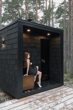 a woman sitting on top of a wooden bench in front of a black shed surrounded by trees