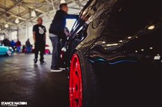 two men standing next to cars in a garage with other people looking at the cars