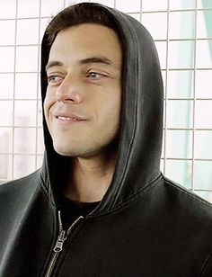 a man wearing a black hoodie in front of a tiled wall