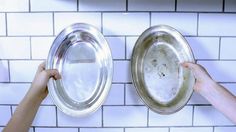 two hands holding silver dishes in front of white tiled backsplashes on a wall