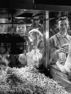 two women in aprons standing behind a glass case filled with popcorn and other food