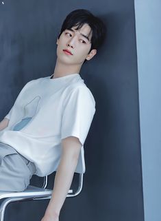 a young man sitting on top of a metal chair next to a black wall and wearing white clothing