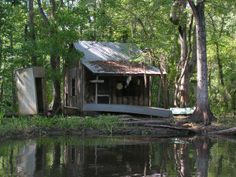 an outhouse sits on the bank of a lake surrounded by trees and grass, with water reflecting it's surface