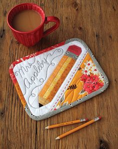 a wooden table topped with a cup of coffee next to a patchwork tray and pencils