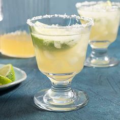 two glasses filled with drinks sitting on top of a blue table covered in sugar and limes