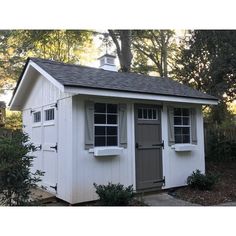 a small white shed with windows and shutters on the side, in front of some trees