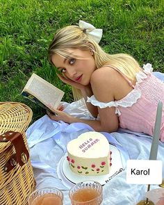 a woman laying on the grass reading a book next to a cake and some drinks