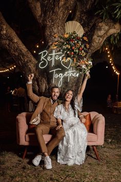 a bride and groom sitting on a pink couch under a tree with their arms in the air