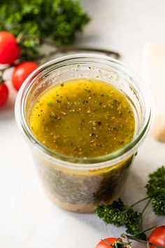 a glass jar filled with olive dressing next to tomatoes and parsley on a white surface