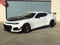 a white chevrolet camaro is parked in front of a garage with an american flag