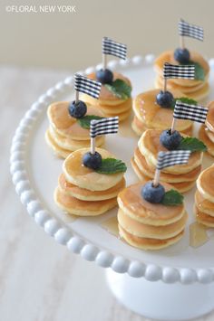 small pancakes with blueberries and flags on them are arranged on a white cake plate