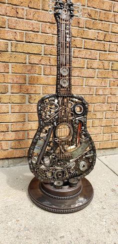 a guitar sculpture sitting on top of a metal stand next to a brick wall with gears all over it