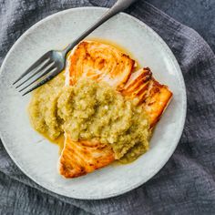 a white plate topped with fish covered in sauce next to a knife and fork on top of a gray cloth