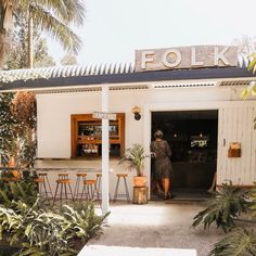 a woman standing in front of a white building with palm trees and plants around it