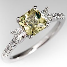 This beautiful yellowish green sapphire ring is centered with one (1) cut-cornered square mixed cut natural sapphire weighing 1.33 carats and set into a four-prong head. The sapphire is flanked to each side by one (1), channel/prong set, square modified brilliant cut diamond. The top face of the shank is accented with five (5), prong set, round brilliant cut diamonds. The ring measures 6.6mm at the top, rises 7.3mm above the finger, tapering to 1.6mm wide and 1.3mm thick at the base of the shank Luxury Green Sapphire Ring Round Cut, Gia Certified Square Cut Sapphire Diamond Ring, Green Sapphire Ring, Yellowish Green, Three Stone Engagement Ring, Stone Engagement Ring, Three Stone Engagement, Three Stone Engagement Rings, Green Sapphire