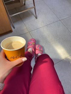 a person with pink shoes holding a cup of coffee in front of their feet on the floor