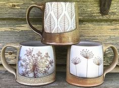 three coffee mugs sitting next to each other on top of a wooden table with leaves painted on them