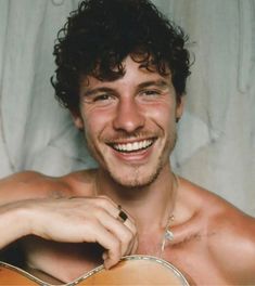 a man with curly hair smiles while holding an acoustic guitar