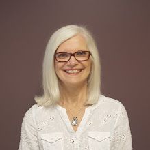 an older woman with white hair and glasses smiles for the camera while standing in front of a brown background